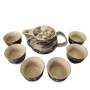 May include: A ceramic tea set featuring a teapot and six matching cups. The teapot and cups have a cream-colored base with a detailed landscape design in shades of gray, brown, and green. The set is ideal for serving tea.