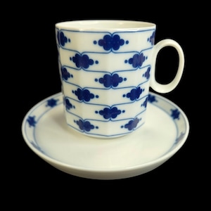 May include: White ceramic teacup and saucer set. The cup has a geometric shape and is decorated with blue cloud-like patterns. The saucer has a matching design. The set is on a black background.