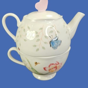 May include: A white ceramic teapot and teacup set. The teapot features a butterfly and floral design with a pink heart-shaped lid handle. The teacup has a bee and floral design. The set is on a blue background.