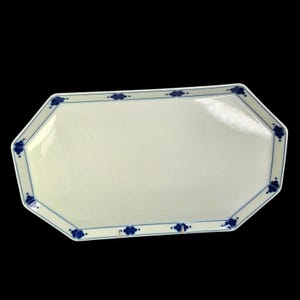 May include: A rectangular, off-white ceramic serving platter with an octagonal shape. The rim is decorated with a blue floral pattern and a thin blue border. The platter is set against a black background.