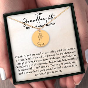 May include: A gold-colored necklace with a round pendant featuring a floral design and the name "Sarah". The necklace is presented in a black box with a message to a granddaughter on her wedding day.