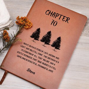 Könnte beinhalten: Ein beigefarbenes Journal in Lederoptik mit dem Text "CHAPTER 70" über drei schwarzen Kiefern-Illustrationen. Unter den Bäumen steht der Text: "YOU'VE GIVEN 70 YEARS OF LOVE, WISDOM, AND LIGHT AND YOU STILL SHINE. THIS BIRTHDAY IS A CELEBRATION OF ALL THAT YOU ARE, AND A GENTLE REMINDER THAT YOUR JOURNEY IS STILL UNFOLDING IN BEAUTIFUL, MEANINGFUL WAYS." Der Name "Steve" ist unten aufgedruckt.