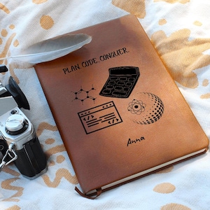 Op de afbeelding: Een bruin leren notitieboek met de tekst "PLAN. CODE. CONQUER" en symbolen voor een rekenmachine, atoom en codering. De naam "Anna" staat onderaan gedrukt. Een witte veer ligt bovenop het notitieboek. Op de achtergrond staat een zilveren camera.
