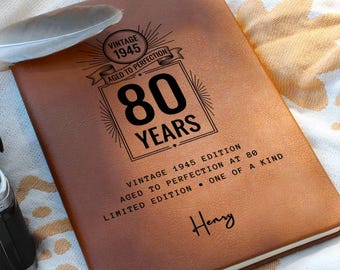 Könnte beinhalten: Ein braunes, ledergebundenes Notizbuch mit dem Text "80 Jahre" in einem dekorativen Rahmen. Der Einband enthält auch den Text "Vintage 1945 Edition" und den Namen "Henry". Eine Feder liegt auf dem Notizbuch.