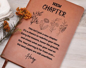 Puede incluir: Un diario de cuero marrón con las palabras "NEW CHAPTER" e ilustraciones florales. El diario tiene texto inspirador y una cinta de marcador. Una ramita de flores naranjas descansa al lado.