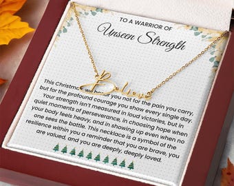 May include: A gold-colored necklace with the word "Believe" in cursive script. The necklace is presented in a gift box with a message that reads "To a Warrior of Unseen Strength". The box is red with a white interior.