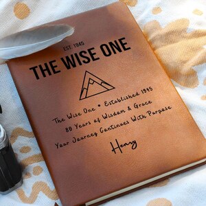 Może przedstawiać: Brązowy skórzany notatnik z czarnym napisem "THE WISE ONE" i "EST. 1945". Notatnik zawiera również grafikę górską i napis "80 Years of Wisdom & Grace". Pióro spoczywa na notatniku.