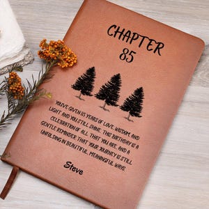 May include: A brown leather journal with the text "CHAPTER 85" and a message about love and wisdom. The journal features three tree illustrations and the name "Steve" at the bottom. A bookmark and floral arrangement are visible.