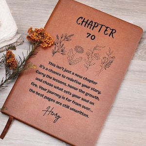 May include: A brown leather journal with the words "CHAPTER 70" and floral illustrations. The journal includes inspirational text and the name "Henry" at the bottom. A sprig of orange flowers rests on the journal.