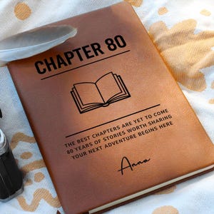 Może przedstawiać: Brązowy, skórzany notatnik z napisem "CHAPTER 80" i ilustracją otwartej książki. Notatnik zawiera napis "THE BEST CHAPTERS ARE YET TO COME" i "YOUR NEXT ADVENTURE BEGINS HERE". Imię "Anna" jest podpisane na dole.