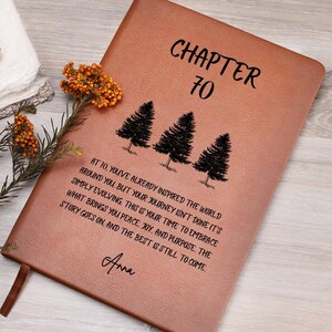 May include: A tan leather journal with the text "CHAPTER 70" and an inspirational message. The journal features three illustrated pine trees and the name "Anna". A decorative floral element is visible. The journal is a thoughtful gift.