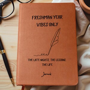 May include: A brown leather-bound journal with the text "FRESHMAN YEAR VIBES ONLY" and "THE LATE NIGHTS, THE LESSONS, THE LIFE" printed in black. A feather illustration and the name "Jamie" are also featured. A pen and glasses are in the background.