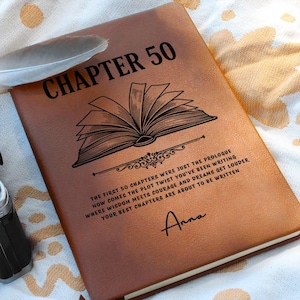 May include: A brown leather journal with the text "CHAPTER 50" and an illustration of an open book. The journal also includes the text "THE FIRST 50 CHAPTERS WERE JUST THE PROLOGUE..." and the name "Anna". A feather rests on the journal.