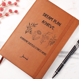 May include: A tan notebook with an elastic closure featuring the black text "DREAM. PLAN. ACHIEVE. ACADEMIC SUCCESS STARTS HERE." and the name "Jamie" alongside a black line art illustration of various wildflowers and a black ballpoint pen.