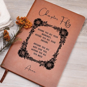 May include: A brown leather journal with the words "Chapter Fifty" in elegant script. The cover features a floral frame with inspirational text. The name "Anna" is inscribed at the bottom. A decorative flower and ribbon are visible.
