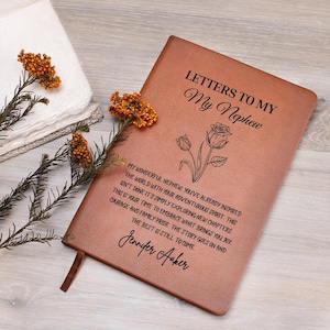 Op de afbeelding: Een bruin leren notitieboek met de tekst "Letters to My Nephew" en een bloemenillustratie. Het notitieboek bevat een persoonlijke boodschap en een lint-bladwijzer. Gedroogde oranje bloemen en een witte doek staan op de achtergrond.