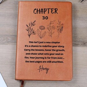 May include: A brown leather journal with the words "CHAPTER 30" and inspirational text. Floral illustrations and the name "Henry" are also featured. The journal has a ribbon bookmark.