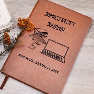 May include: A brown leather journal with the text "Jamie's Reset Journal" and the words "Refocus. Rebuild. Rise." The journal features illustrations of books, a coffee cup, and a laptop. A brown ribbon bookmark is visible.