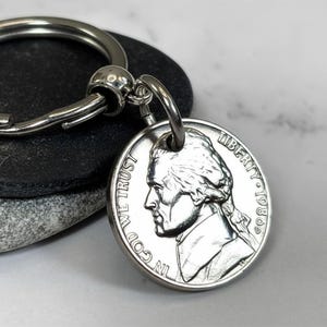 May include: A silver-toned metal keychain featuring a circular split ring attached to a 1980 United States nickel coin. The coin displays the profile of Thomas Jefferson, the text IN GOD WE TRUST, the word LIBERTY, and the year 1980.