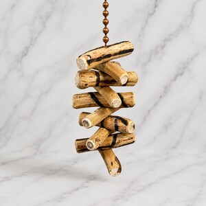 May include: A ceiling fan pull chain featuring a series of small, cylindrical wooden segments arranged in a vertical, staggered formation. Each light brown wooden piece displays dark, charred-looking horizontal markings and a natural grain texture.