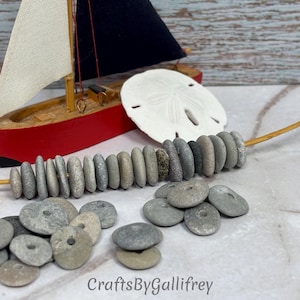 May include: A collection of small, flat, gray beach stones with drilled center holes, some strung on a thin wooden twig. A miniature red and white sailboat model and a white sand dollar are positioned behind the stones. Text reads CraftsByGallifrey.