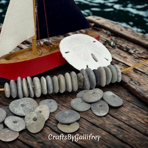 May include: A miniature wooden sailboat with a red hull and a white sail, alongside a sand dollar and a collection of gray stones. The scene is set on a weathered wooden surface, evoking a nautical theme.