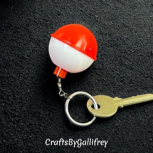 May include: A novelty keychain featuring a red and white fishing bobber design. The bobber is attached to a silver keyring with a gold-colored key. The keychain is set against a dark textured background.