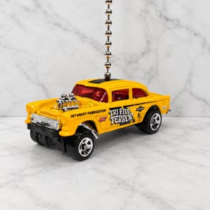 May include: A yellow toy car ceiling fan pull chain featuring a black stripe on the roof, red windows, and a silver engine block. The side displays the text VETUSKEY FABRICATION, 427, TRI FIVE TERROR, and a Chevrolet logo on a black background.