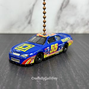 May include: A blue and yellow race car ornament with the number 55 on the side. The car has sponsor logos and the words "Monte Carlo" on the front. The ornament is suspended by a chain.