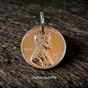 May include: A copper-colored 1972 Lincoln penny coin pendant featuring a silver-toned metal jump ring attached at the top. The coin displays the profile of Abraham Lincoln, the text IN GOD WE TRUST, LIBERTY, and the year 1972 with a D mint mark.