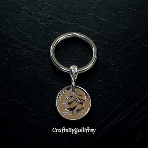 May include: A silver-colored keychain with a dime pendant. The dime features a profile of a person and the word "LIBERTY". The keychain is attached to a silver ring. The background is black. The text "CraftsByGallifrey" is at the bottom.