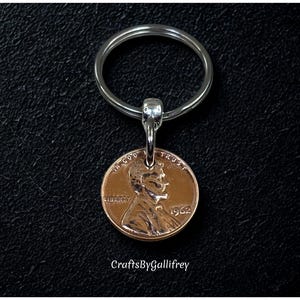 May include: A copper penny keychain featuring a 1962 penny. The penny is attached to a silver-colored metal keyring. The keychain is displayed against a black background. The text "CraftsByGallifrey" is in the lower right corner.