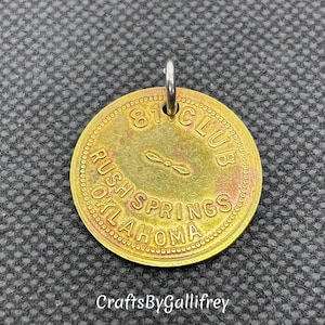 May include: A gold-colored metal token with the text "81 Club Rush Springs Oklahoma" and a small propeller design. The token has a hole at the top for hanging.
