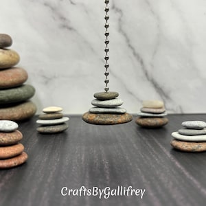 May include: A collection of stacked stones in various sizes and colors, arranged in balanced formations. Some stacks are on a dark surface, while one is suspended from a chain. The image includes the text "CraftsByGallifrey".