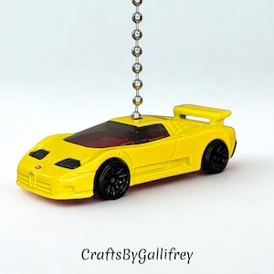 May include: A bright yellow toy car with black wheels and a rear spoiler, suspended by a silver chain. The car has a red interior and a sleek design. The text "CraftsByGallifrey" is at the bottom.
