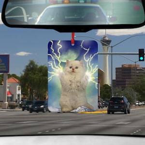 Könnte beinhalten: Eine weiße Katze mit blauen Augen ist von Blitzen auf einem blauen Hintergrund umgeben. Die Katze steht auf einer weißen Wolke. Das Bild ist auf einem Auto-Lufterfrischer.