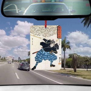 May include: Rectangular car air freshener featuring a Japanese woodblock print of a samurai in action. The samurai is depicted in black and blue clothing, wielding a sword. The background includes Japanese text and a red accent.