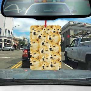 May include: A car air freshener with a pattern of many Shiba Inu dog faces. The air freshener is hanging from the rearview mirror of a car. The car is driving down a street with other cars and buildings in the background.