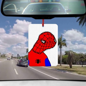 Könnte beinhalten: Ein weißer Auto-Lufterfrischer mit einem roten und blauen Cartoon-Bild von Spider-Man mit einer roten Spinne auf der Brust. Der Lufterfrischer hängt an einem Rückspiegel in einem Auto.