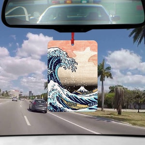 Könnte beinhalten: Ein Auto-Lufterfrischer mit einem farbenfrohen Druck einer großen Welle, die gegen einen Berg kracht. Die Welle ist blau, weiß und grün, und der Berg ist hellbraun. Der Hintergrund ist hellrosa.