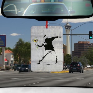 Könnte beinhalten: Ein Auto-Lufterfrischer mit einer schwarz-weißen Schablone einer Person, die einen Blumenstrauß wirft. Der Hintergrund ist eine strukturierte graue Oberfläche.