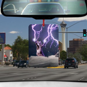 Könnte beinhalten: Ein Auto-Lufterfrischer mit einer lila Katze, die Blitze vor einem stürmischen Himmel hält. Die Katze steht auf einer Wolke.