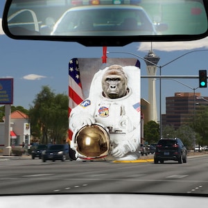 May include: A car air freshener featuring a gorilla wearing an astronaut suit with a gold helmet. The gorilla is in front of an American flag.