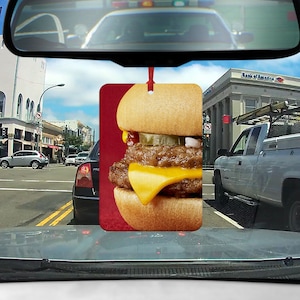 May include: A car air freshener featuring a close-up of a cheeseburger with lettuce, tomato, and a pickle on a sesame seed bun. The air freshener is red and white.
