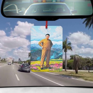 Pode incluir: Um aromatizador de carro com um retrato de um homem de terno marrom em pé em um campo de flores com montanhas ao fundo. O aromatizador está pendurado no espelho retrovisor de um carro.