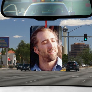 May include: A car air freshener featuring a close-up photo of Nicolas Cage with his eyes closed. The photo is set against a blurry background of a city street with a tall building and a blue sky.