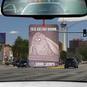 May include: A car air freshener featuring a white moth with large eyes and the text "Ya'll got any uhhh... ...uhhh...lamps" on a pink background.