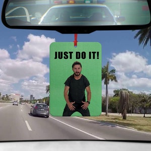 Könnte beinhalten: Rechteckiger Lufterfrischer mit dem Text "JUST DO IT!" und einem Bild eines Mannes in schwarzem Hemd und Hose. Der Lufterfrischer hängt an einer roten Schnur vor der Windschutzscheibe eines Autos, mit Blick auf eine Autobahn und Palmen.