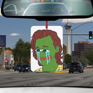 Könnte beinhalten: Eine grüne Comicfigur mit braunen Haaren weint blaue Tränen. Die Figur trägt ein gelbes und braunes Karohemd. Das Bild ist auf einem weißen Auto-Lufterfrischer.