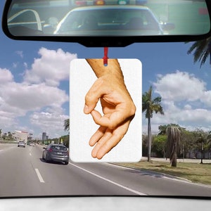 May include: A white car air freshener with a hand making the "okay" gesture. The hand is in focus and the background is blurred.